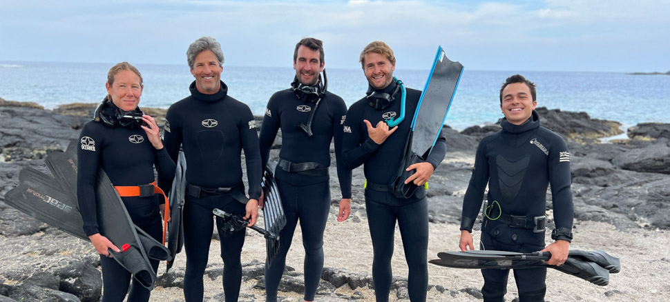 A group of people stand side by side wearing wetsuits and holding freediving long fins on a lava rock beach in Hawaii