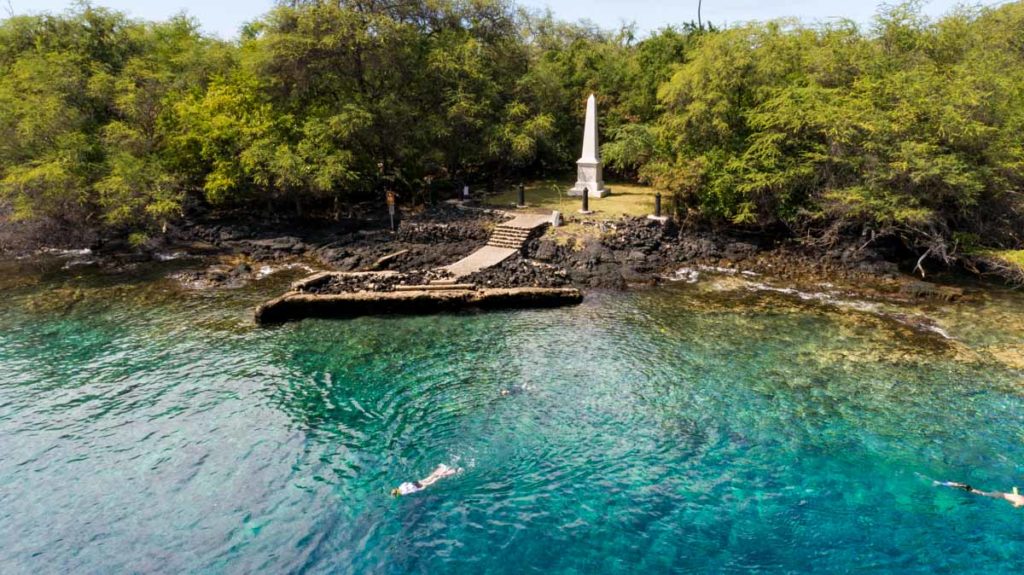snorkelers in the water near shore with the captain cook obelisk monument in background with lots of trees and rocky coast