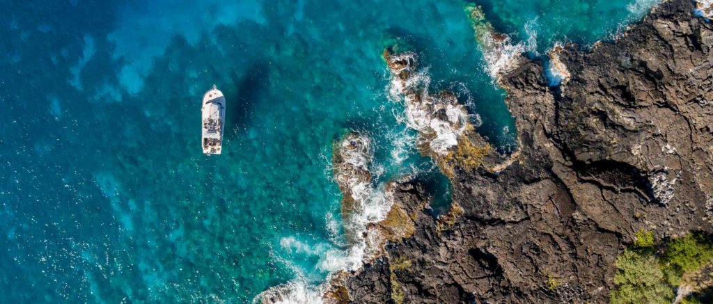 overhead aerial view of a boat floating in the ocean with reef below near a rocky lava coast