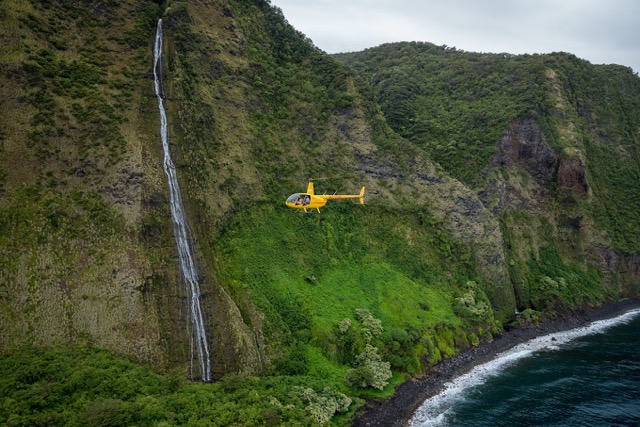 helicopter flying past a waterfall coming from a lush valley with black sand below