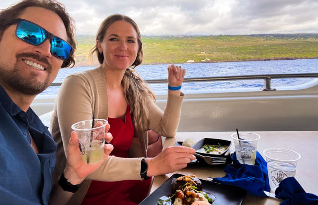 2 people sit together at a table with food and drinks on a boat. The Kona coastline is in the background