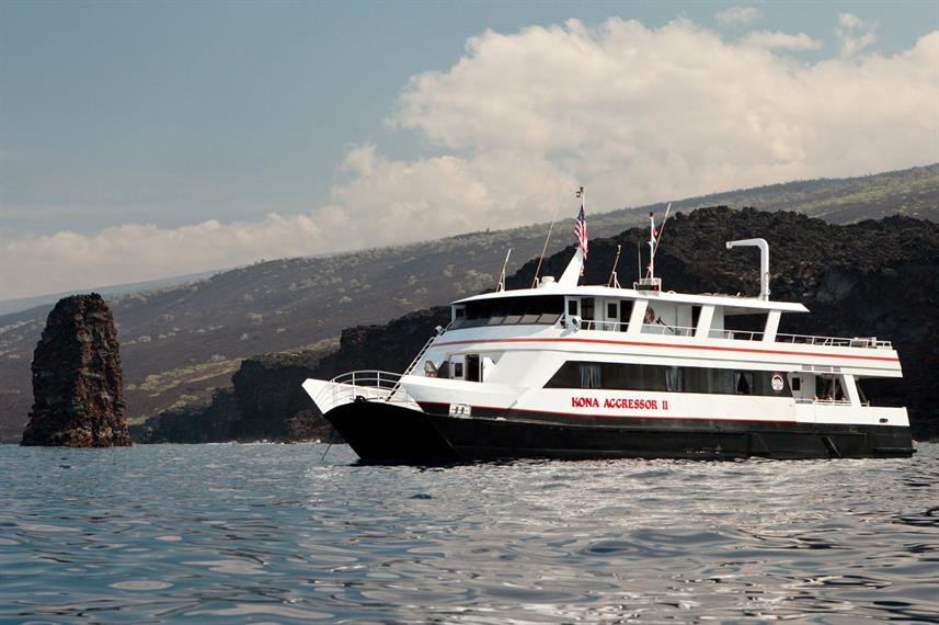 A large catamaran sits offshore near a rock pinnacle with rough lava rock shoreline behind