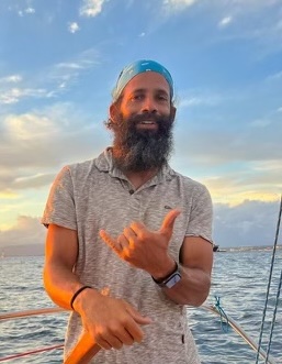 man with bandana and beard holding a sailboat tiller with sunset behind