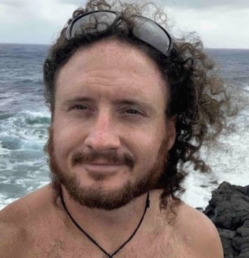 headshot of a man with curly red hair held back by suinglasses standing in front of a rough ocean and lava rock below