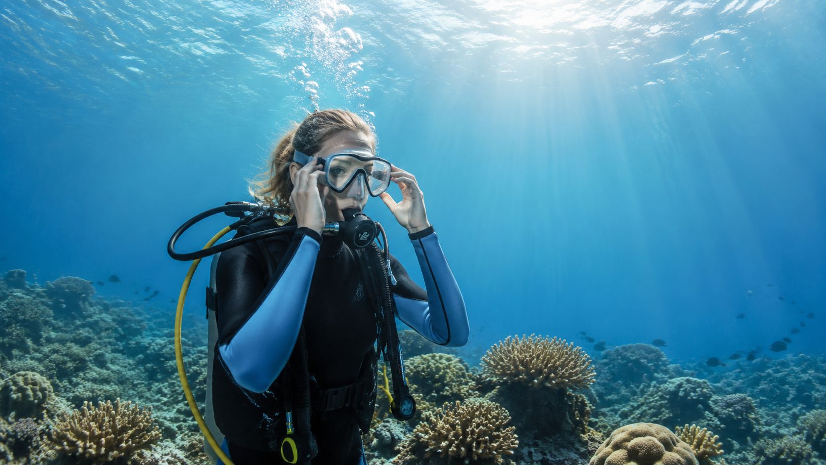 A female scuba diver adjusting her mask while swimming over a colorful coral reef in clear blue water.