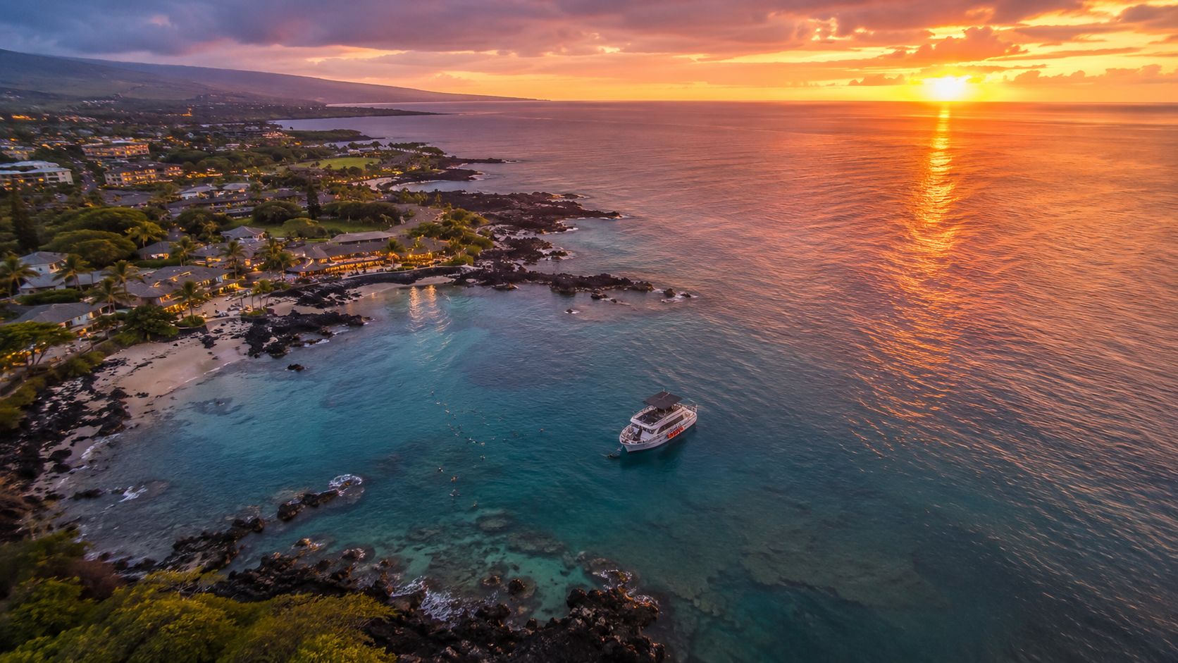 An aerial view of a tropical Hawaiian coastline at sunset with a boat in the turquoise water.