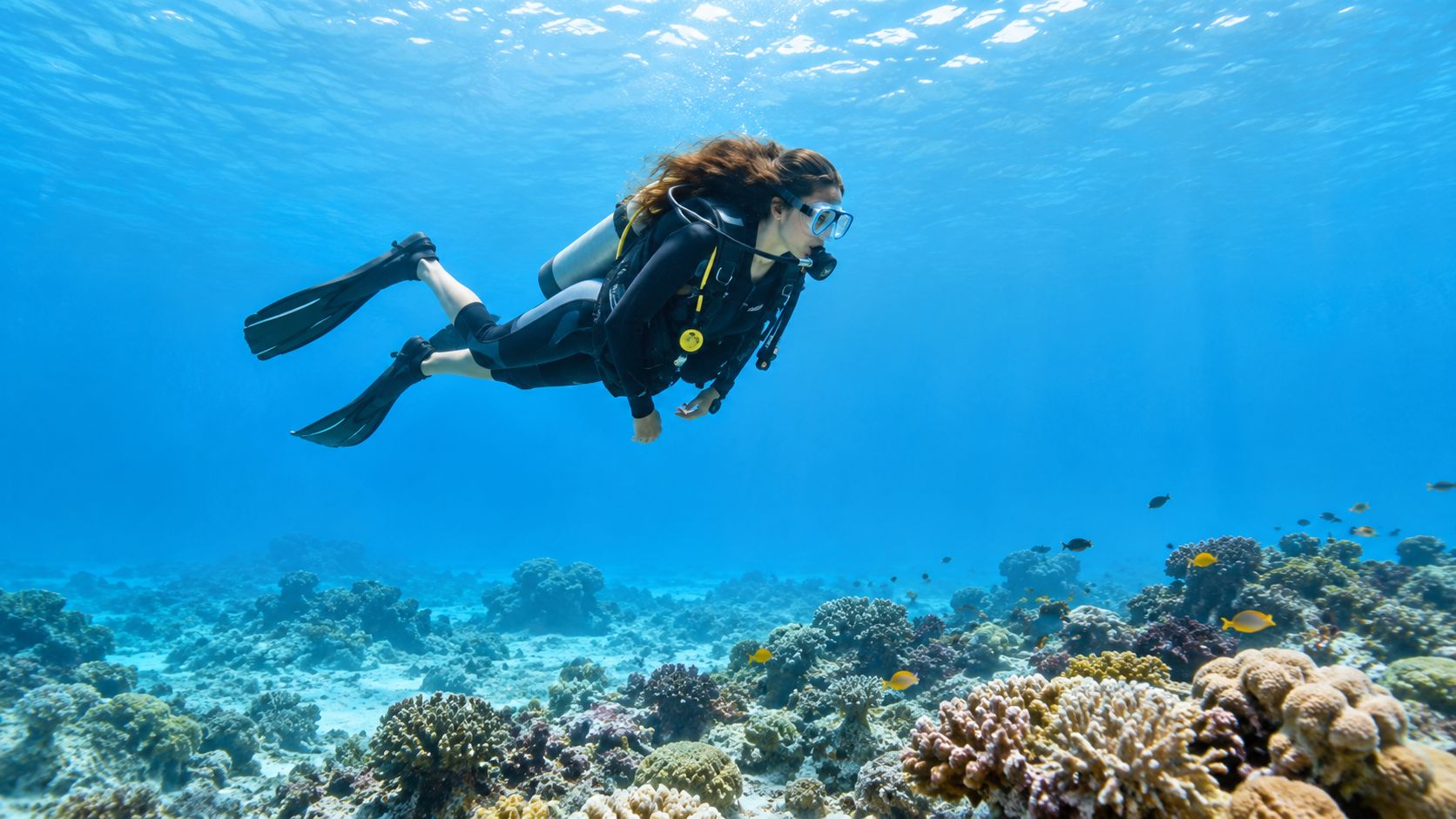 A female scuba diver explores a vibrant coral reef filled with tropical fish in clear blue water.