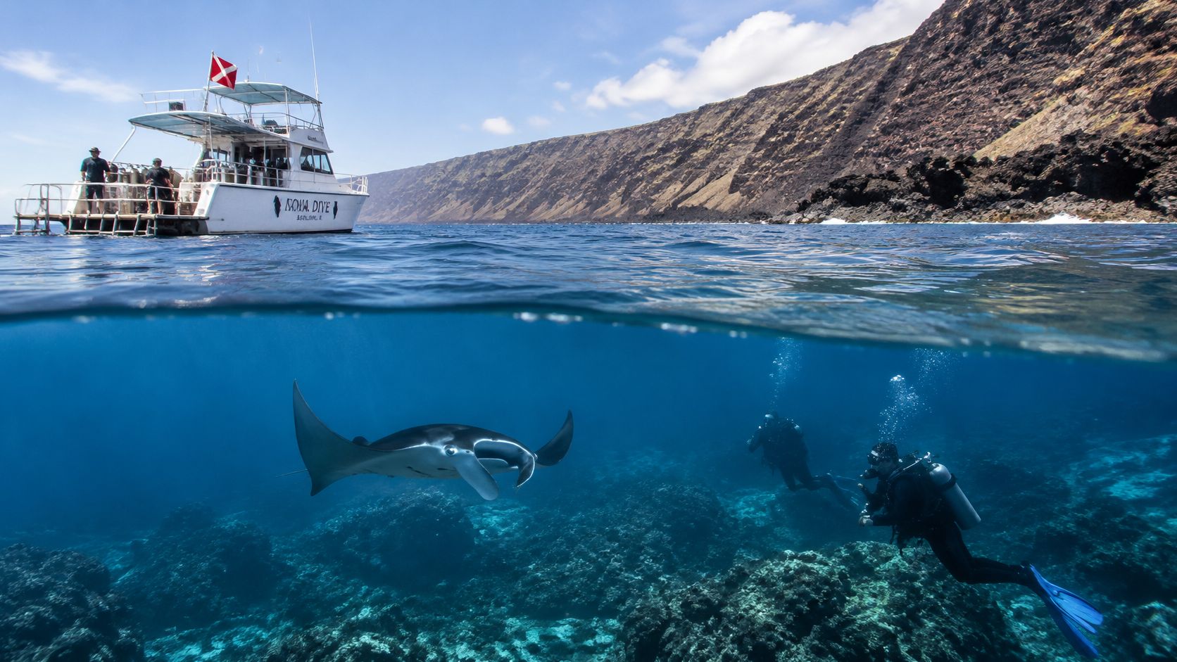 A boat floats above divers swimming alongside a graceful manta ray in clear tropical ocean waters.