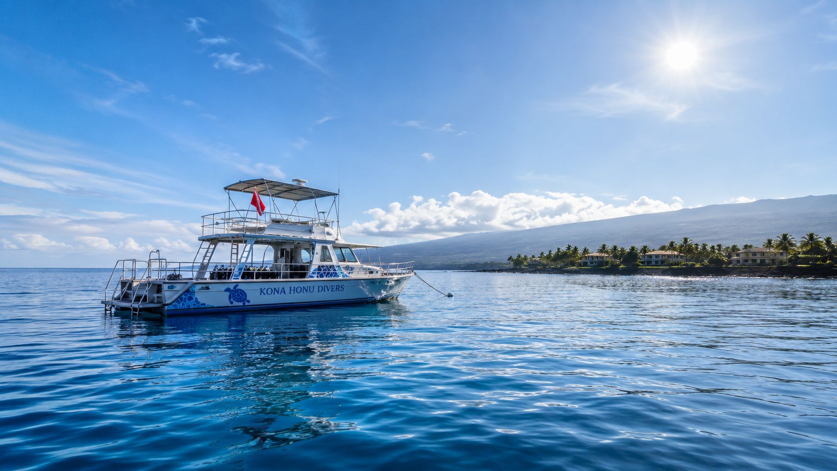 A Kona Honu Divers scuba diving boat anchored in calm tropical waters near a coastal island landscape.