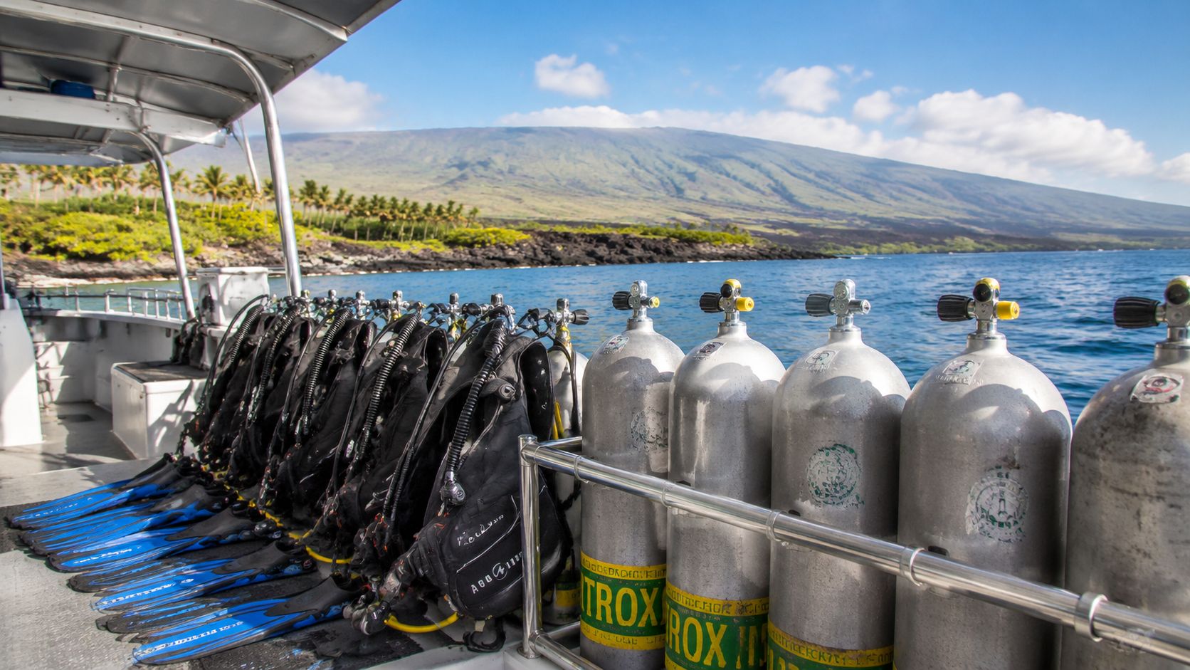 A row of scuba diving tanks and blue fins lined up on a boat near a tropical island.
