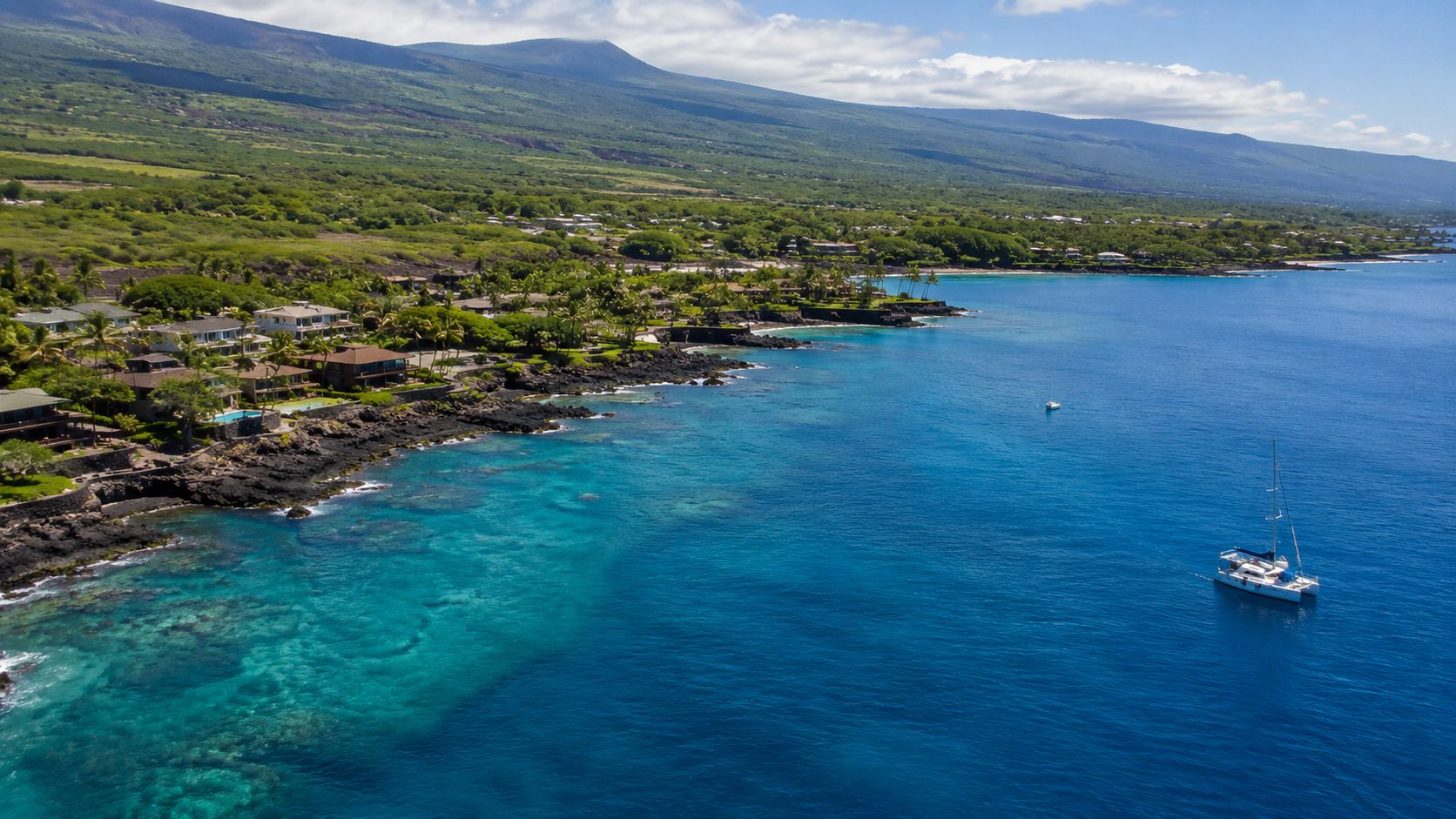 An aerial view of a coastline in Hawaii with a luxury catamaran sailing on clear blue water