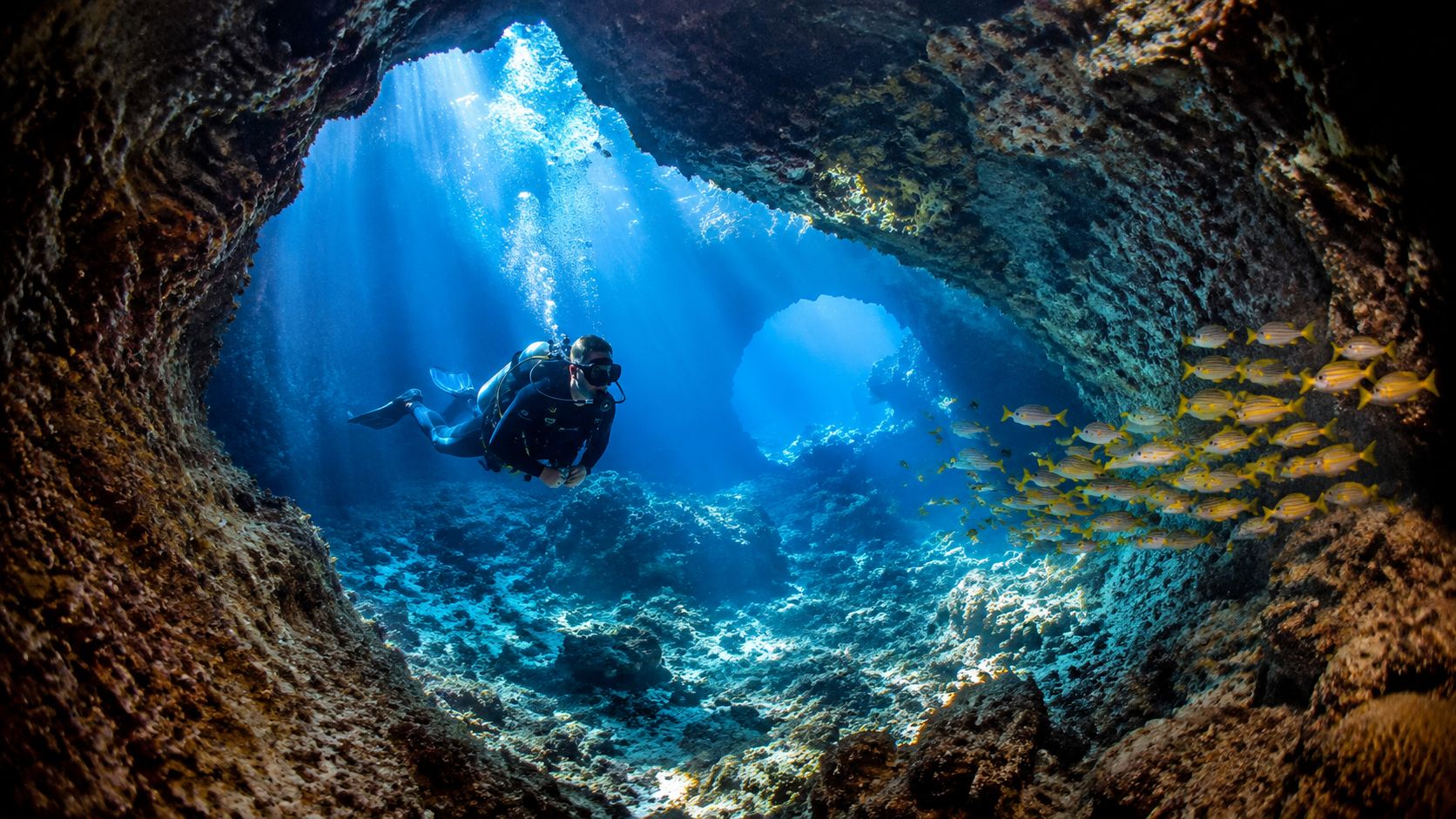 A scuba diver explores a stunning underwater volcanic cave formation illuminated by bright beams of sunlight.