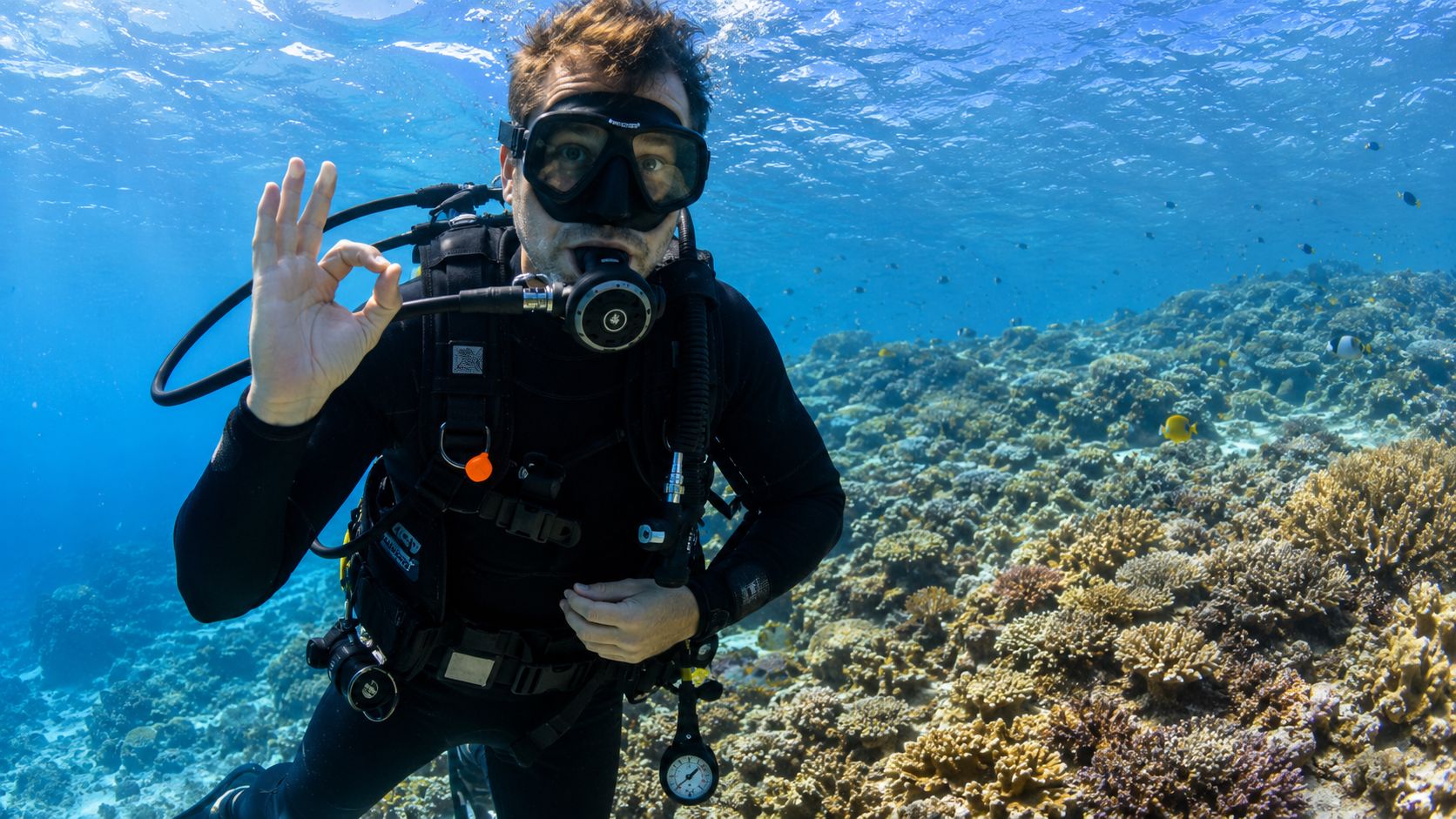A scuba diver making an okay gesture while swimming over a vibrant coral reef in clear blue water.