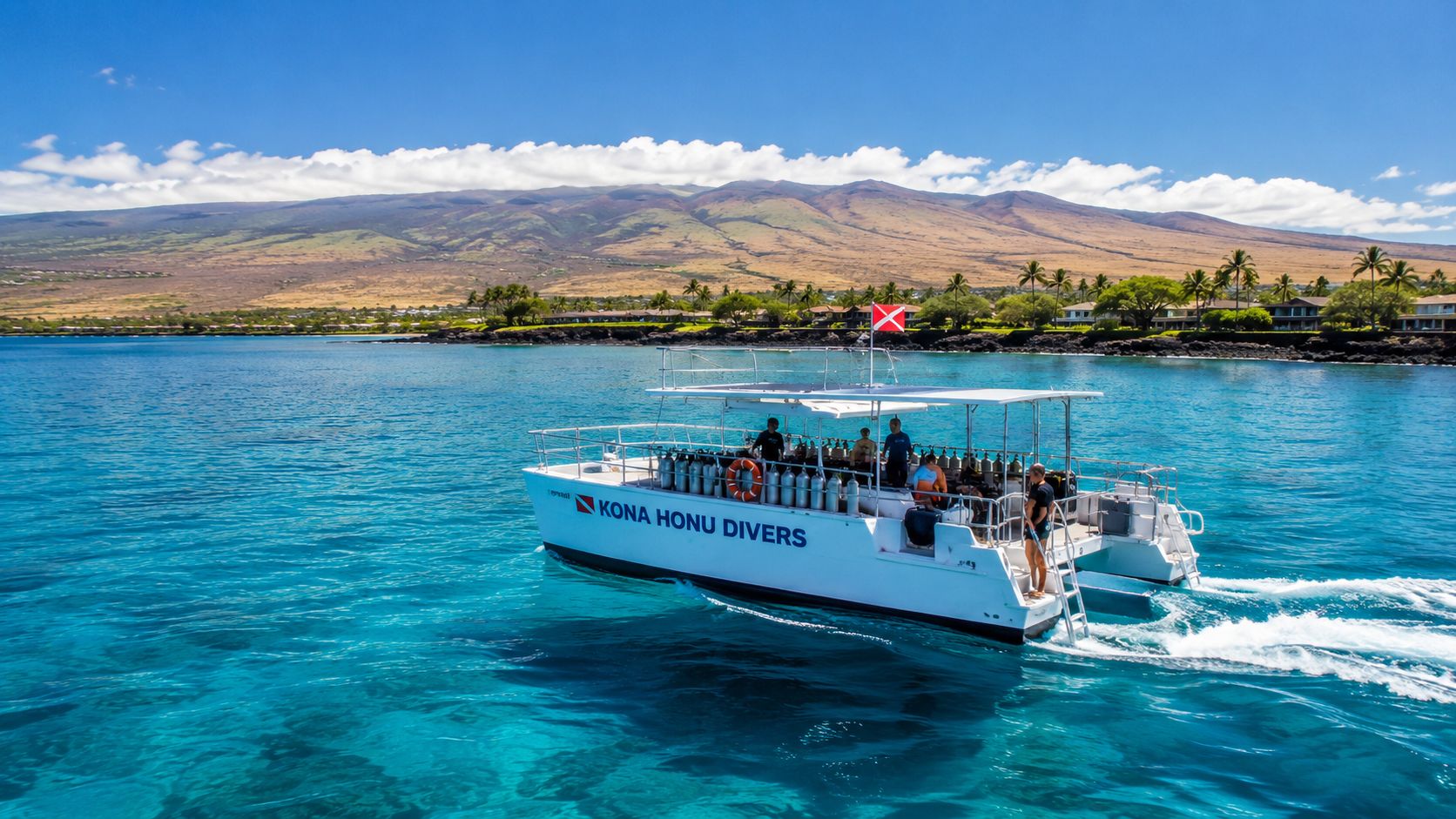 A white catamaran boat labeled Kona Honu Divers cruising across bright blue ocean waters near a mountain coastline.