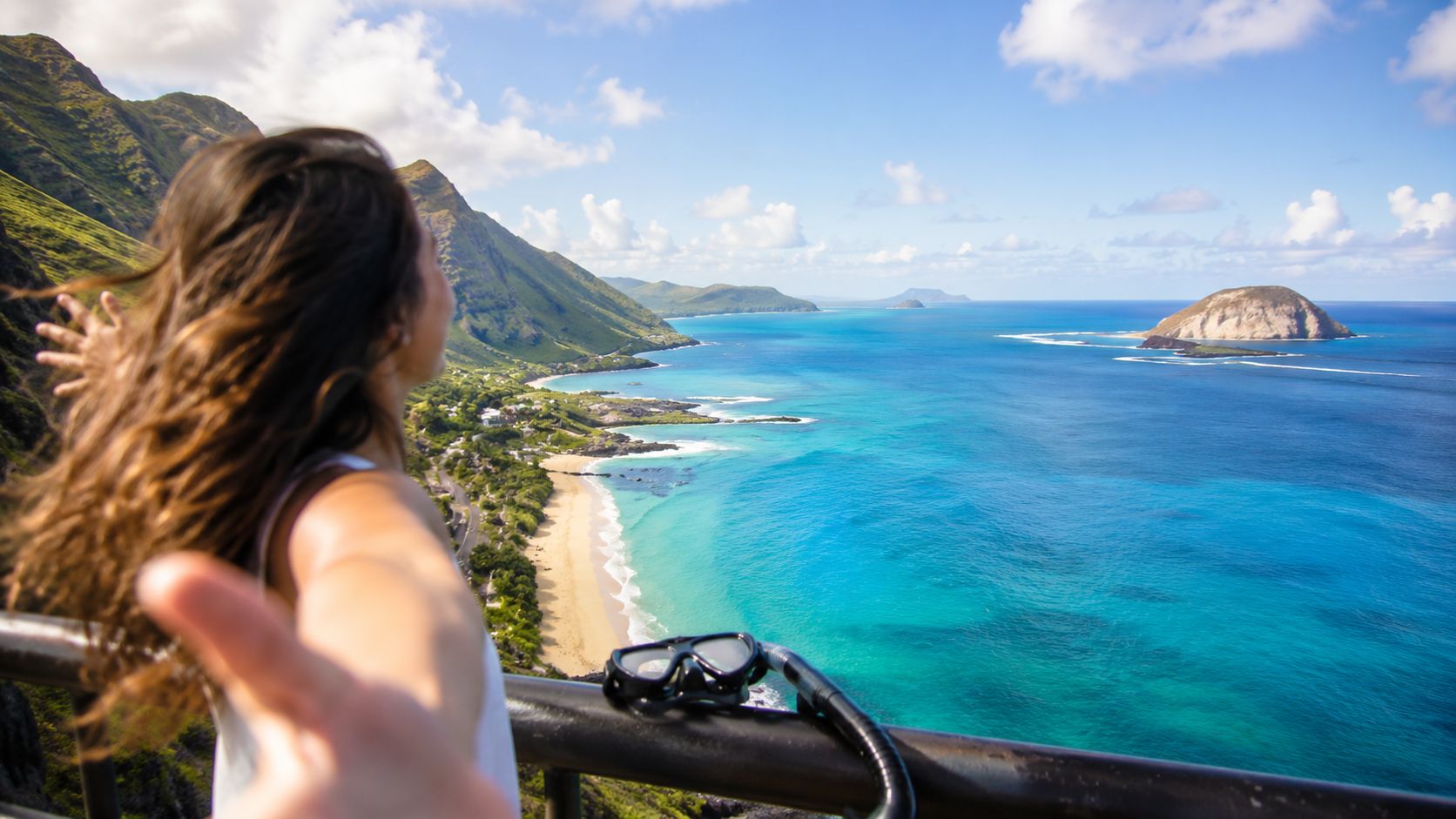 A woman overlooking a scenic Hawaiian coastline with turquoise water, mountains, and a snorkeling mask nearby.