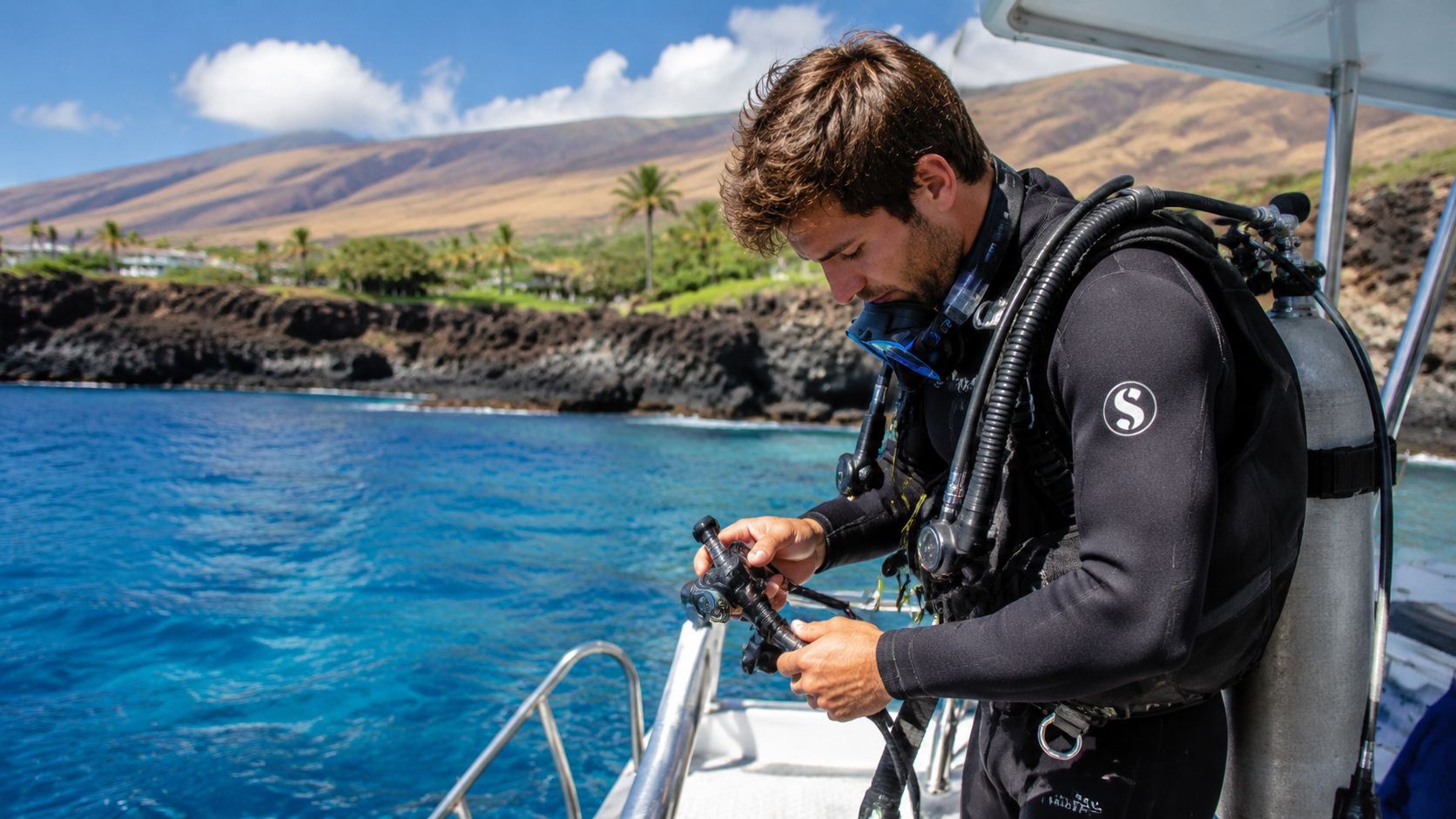 A scuba diver wearing a black wetsuit prepares his gear on a boat before diving in Hawaii.