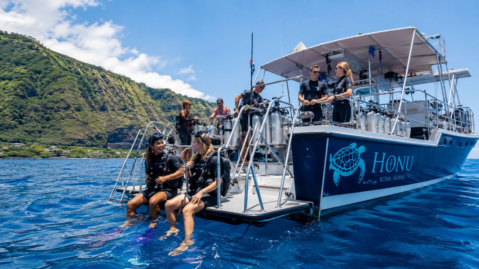 Scuba divers sitting on the back deck of a dive boat in Kona, Hawaii with scenic cliffs behind.
