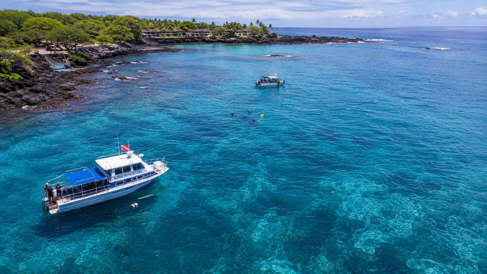 A Kona Honu Divers boat anchored in clear blue Hawaiian waters with snorkelers swimming nearby in the ocean.