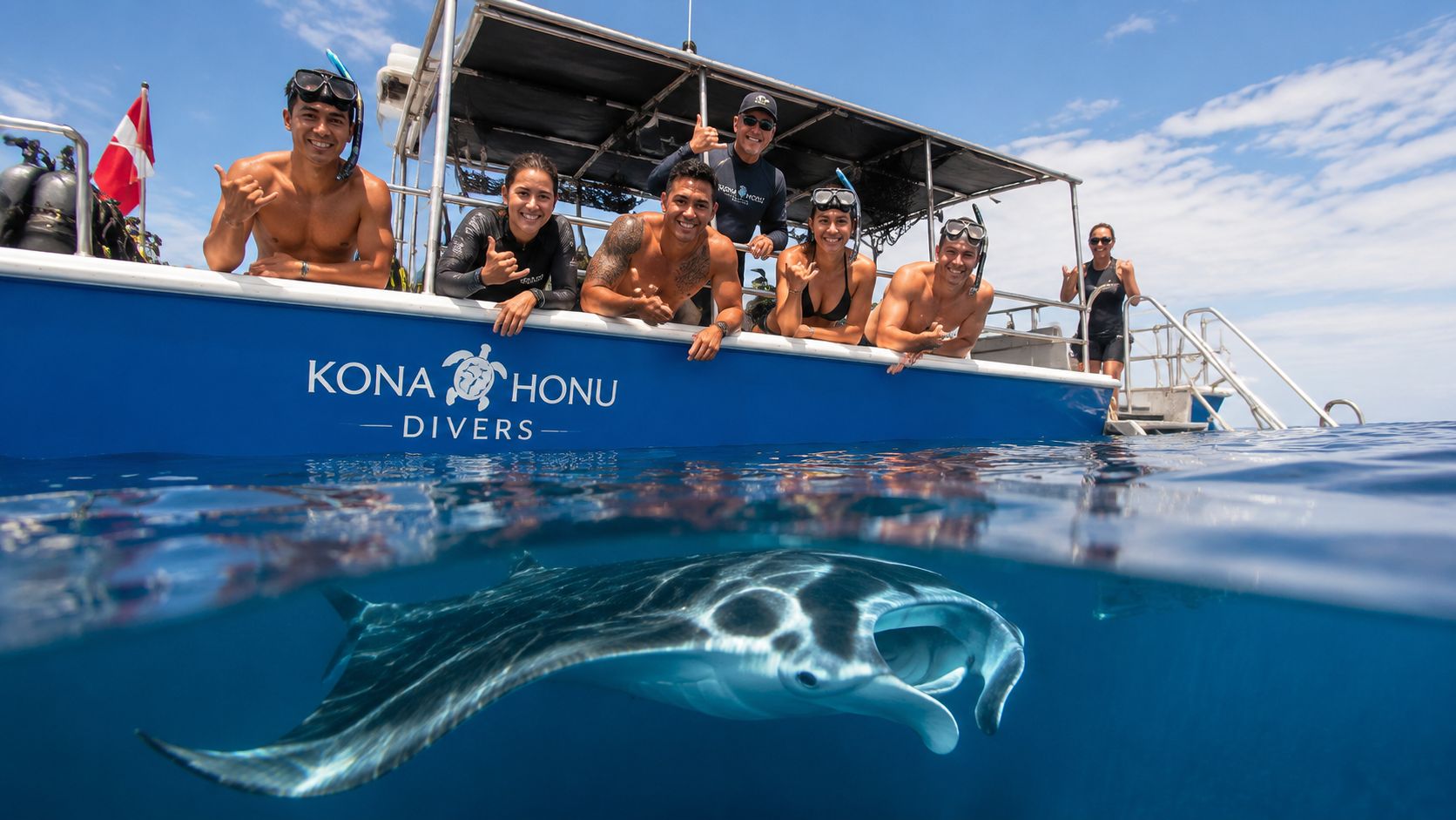 A group of snorkelers and divers smile from a boat above a majestic manta ray swimming underwater.