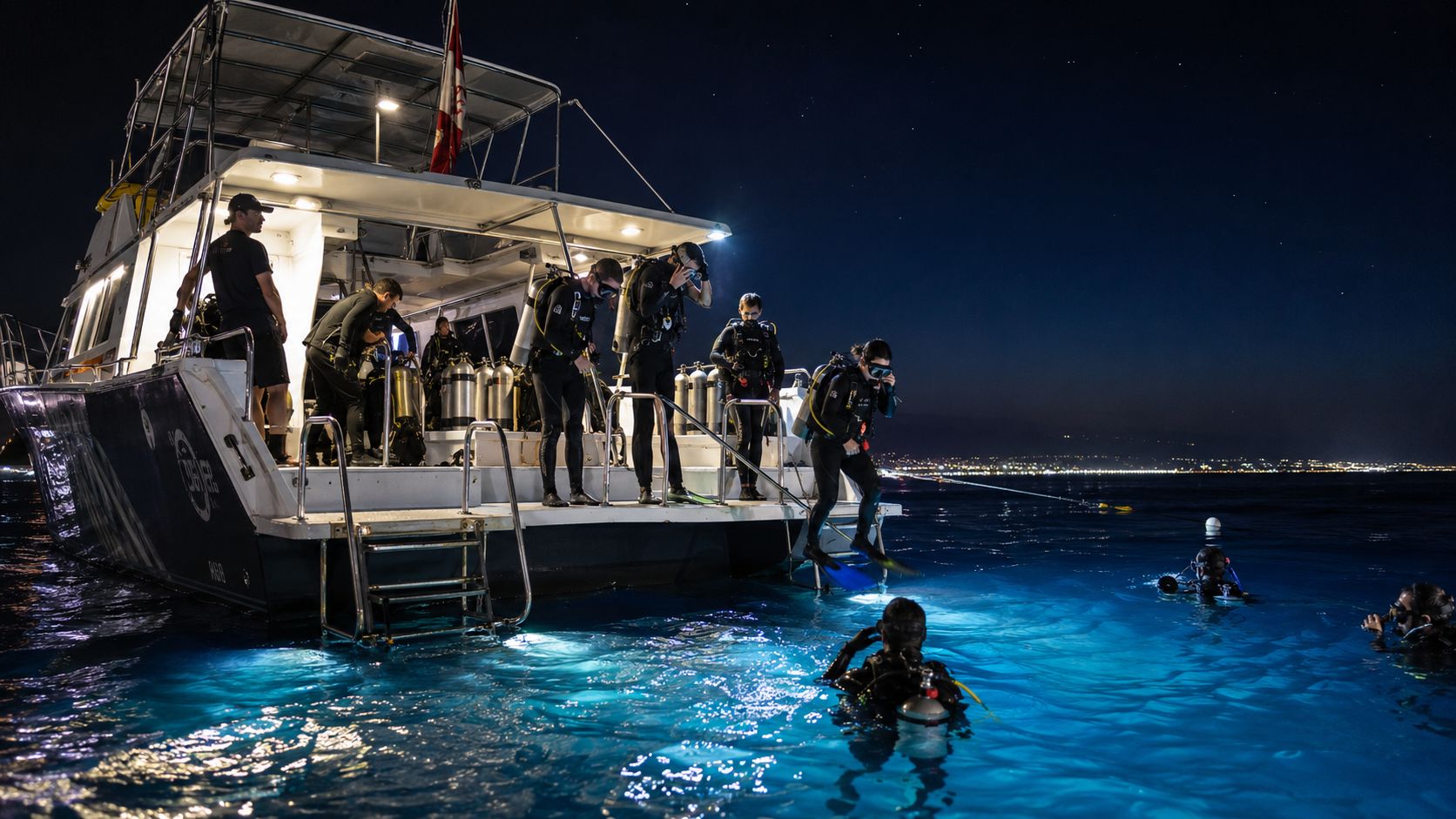 Scuba divers preparing to enter the ocean from a boat for a night dive under stars.