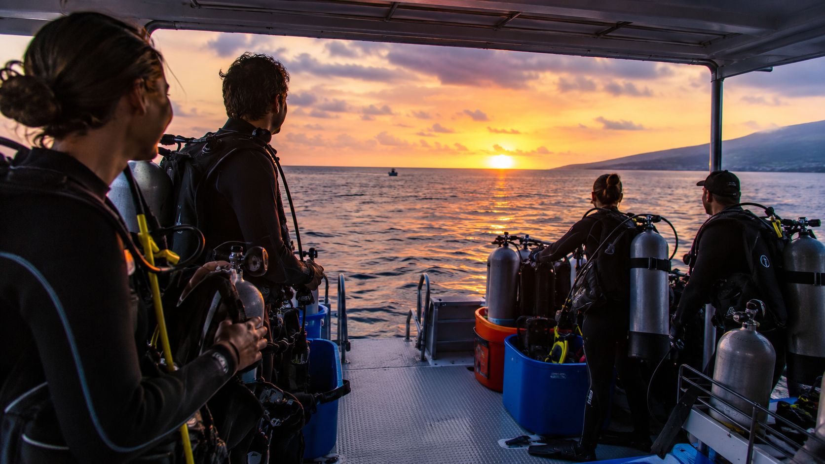 A group of scuba divers on a boat watch a beautiful sunset over the ocean.