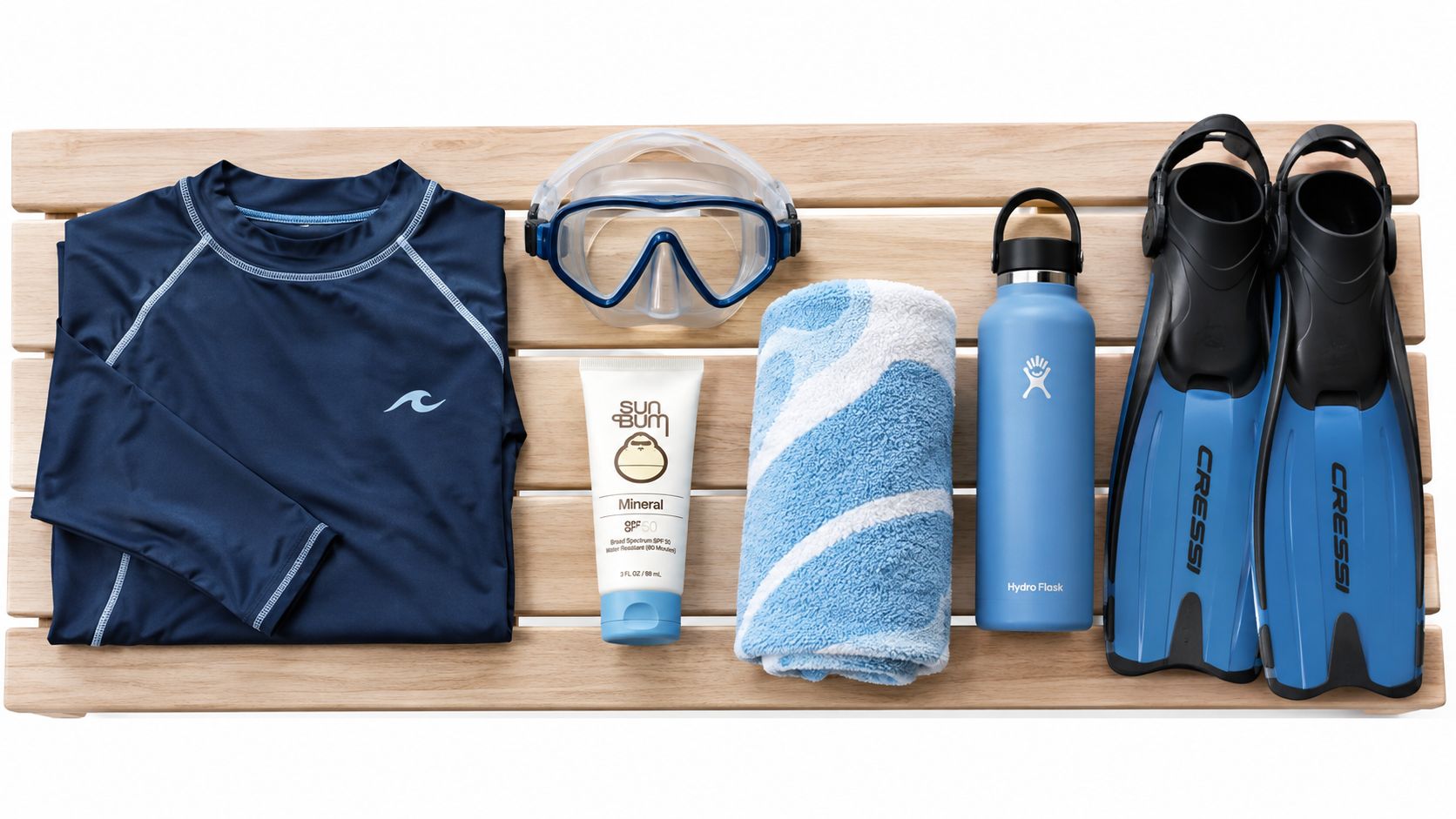 A flatlay of snorkeling gear on a wooden bench, including a rash guard, mask, sunscreen, towel, bottle, and fins.