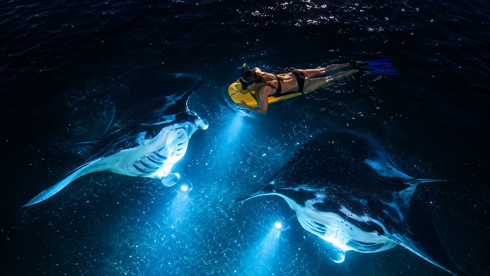 A swimmer with a snorkel and board encountering two large manta rays in dark blue water.