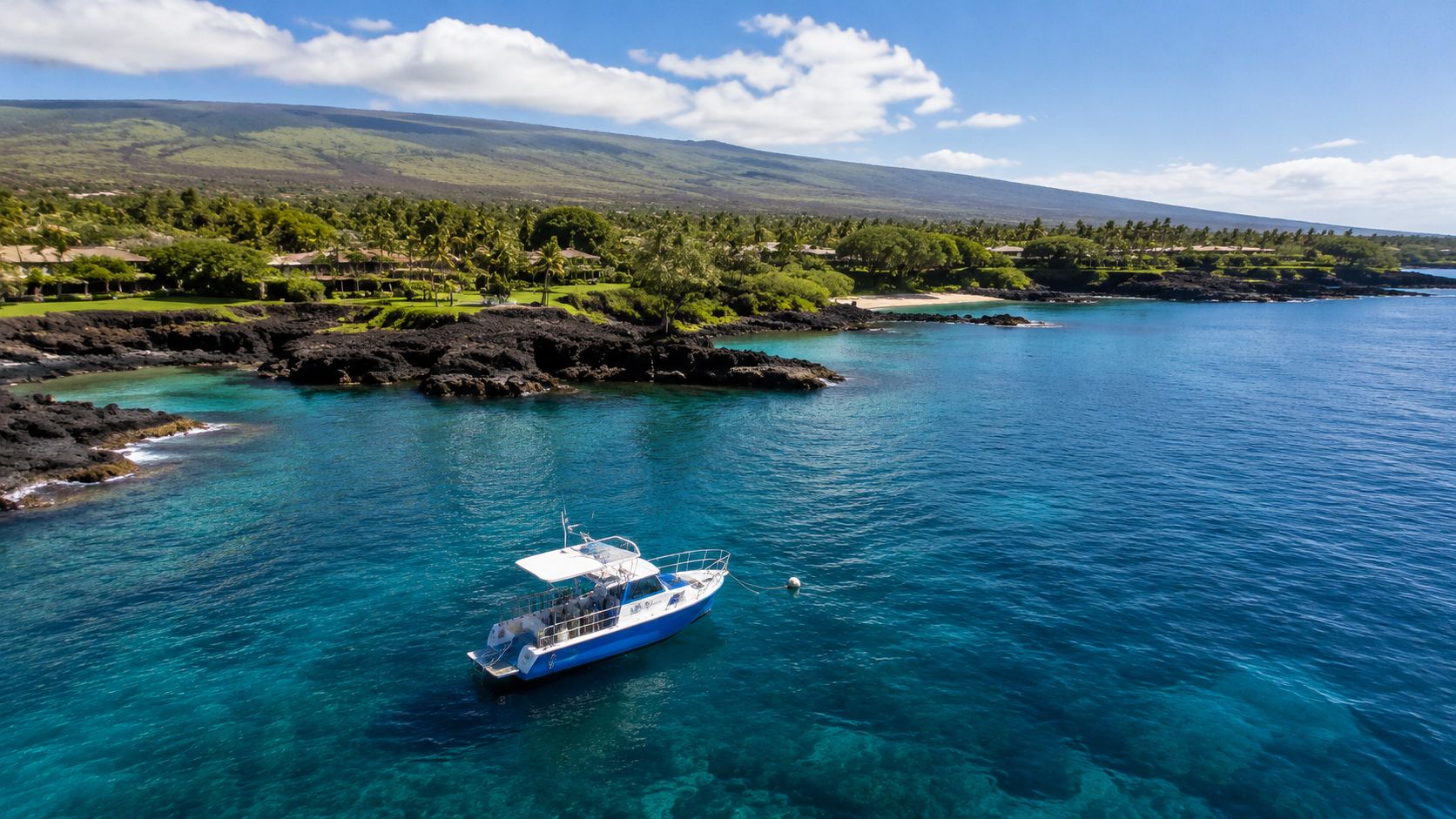 A small tour boat floating in crystal clear blue tropical water near a lush volcanic coastline.