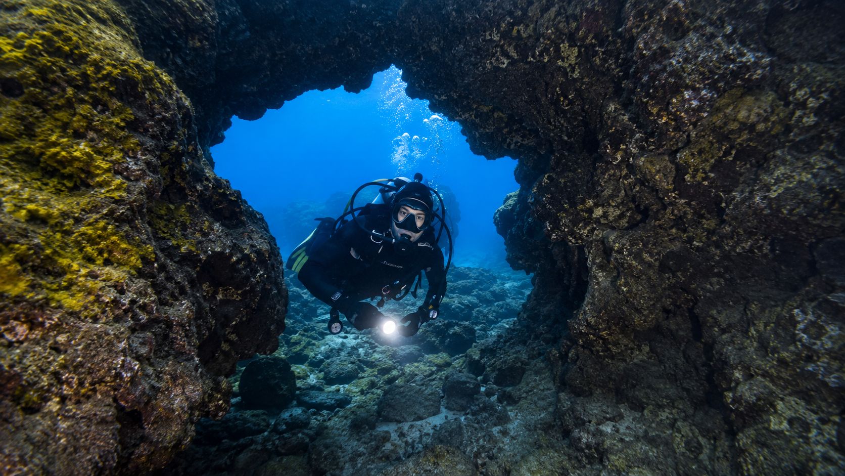 A scuba diver explores a rocky underwater cave surrounded by colorful coral reefs in deep blue water.